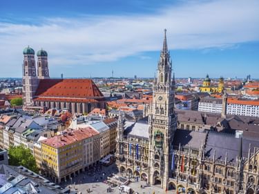 Luftaufnahme der Münchner Innenstadt mit den Zwillingstürmen der Frauenkirche mit rotem Dach und dem verzierten Turm des Neuen Rathauses am Marienplatz.