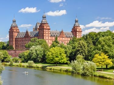 Rotes Sandsteinschloss Aschaffenburg mit drei Türmen hinter grünem Park und Fluss. Zwei Schwäne schwimmen im Wasser unter blauem Himmel.