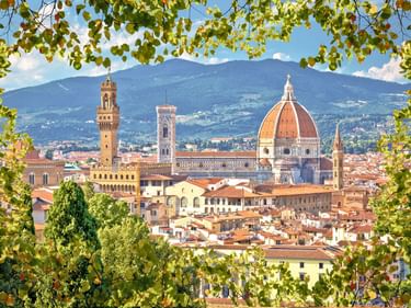 Florenz Stadtansicht umrahmt von grünen Blättern, zeigt die rote Domkuppel, Glockentürme und Terrakotta-Dächer mit Bergen im Hintergrund.