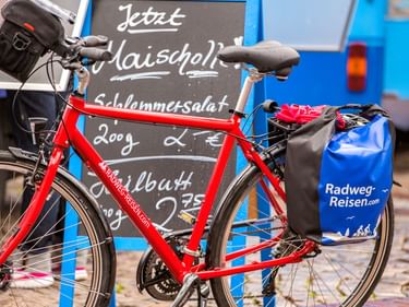 Rotes Reiserad mit blauen Radweg-Reisen.com Packtaschen vor einer Kreidetafel mit Angeboten für Schommersalat und Grillbutt.