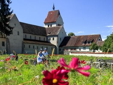 Two cyclists riding past colorful flower garden with historic white church and traditional buildings on Reichenau Island under blue sky.