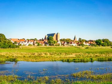 Blick auf die Stadt Verden mit Kirchtürmen und traditionellen Häusern über grüne Wiesen und die Weser unter klarem blauen Himmel.