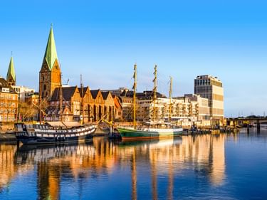 Historische Segelschiffe im Bremer Hafen mit Kirchtürmen und modernen Gebäuden, die sich im ruhigen Wasser unter blauem Himmel spiegeln.