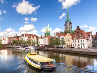 Gelbes Ausflugsboot auf der Trave mit historischer Lübecker Altstadt, bunten Gebäuden und Kirchtürmen unter blauem Himmel mit weißen Wolken.