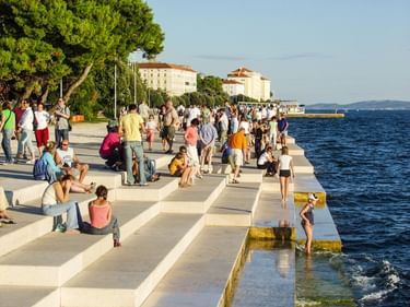 Zadar Uferpromenade mit Menschen, die auf weißen Stufen sitzen. Historische Gebäude und Bäume säumen den Weg entlang der Adria.