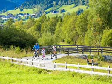 Drei Radfahrer am Murradweg mit weißem Holzzaun, grünen Wiesen und Alpendorf im Hintergrund unter blauem Himmel.