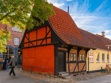 Bunte Fachwerkhäuser in der Altstadt von Køge mit roten und gelben Fassaden, roten Ziegeldächern und Kopfsteinpflaster. Fußgänger gehen an den Gebäuden vorbei.