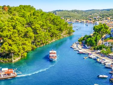Luftaufnahme des Hafens von Gaios auf Paxos mit zwei Ausflugsschiffen in einem schmalen Kanal, gesäumt von grünen Bäumen und Häusern.