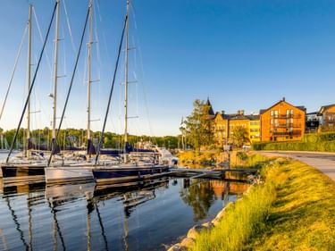 Segelboote im Yachthafen von Gustavsberg mit gelben Gebäuden und grünem Uferweg im Abendlicht.