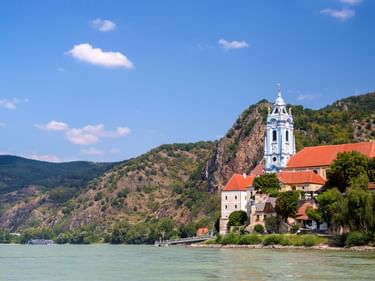 Dürnstein mit blauem Barockkirchturm an der Donau, umgeben von felsigen Hügeln und Weinbergen unter blauem Himmel.