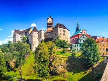 Mittelalterliche Burg Loket auf einem Felsenhügel über bunten historischen Gebäuden. Besucher gehen über eine Brücke mit grünen Bäumen und Fluss darunter.