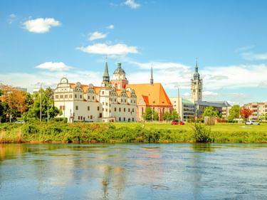 Dessau Palace with white facade and towers beside the Elbe River, with church featuring orange roof and green riverbank under blue sky.