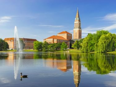 Kieler Rathausturm mit rotgedeckten Gebäuden spiegelt sich in einem ruhigen Teich. Links spritzt ein Springbrunnen, grüne Bäume säumen das Ufer.