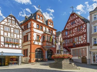 Bunte Fachwerkhäuser umgeben einen Brunnen auf dem Altstadtplatz von Bernkastel-Kues unter blauem Himmel mit weißen Wolken.