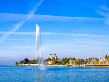 Bodenseeufer in Friedrichshafen mit hoher Fontäne, Schlosskirche mit zwei Türmen, Yachthafen mit Segelbooten und blauem Himmel.
