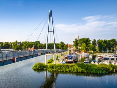 Modern cable-stayed pedestrian bridge spanning a waterway in Gizycko marina. Moored boats and green vegetation visible along the shore under blue sky.