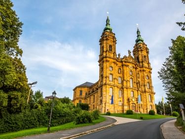 Gelbe Barockbasilika mit Zwillingstürmen und grünen Kuppeln in Bayern, eingerahmt von Bäumen entlang einer Kurvenstraße unter blauem Himmel.
