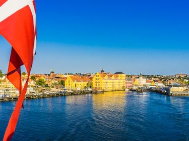 Blick auf den Hafen von Svendborg mit bunten historischen Gebäuden entlang der Uferpromenade. Eine rot-weiße dänische Flagge ist im Vordergrund sichtbar.