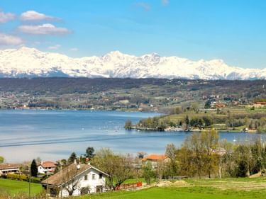 Lago di Viverone mit grünen Wiesen und Häusern im Vordergrund, ruhigem blauen See und schneebedeckten Alpengipfeln unter klarem Himmel.