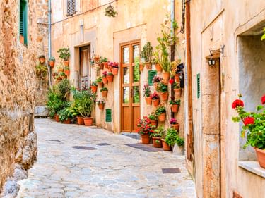 Enge Kopfsteinpflastergasse in Valldemossa mit verwitterten Steinmauern, Holztür und zahlreichen Terrakotta-Blumentöpfen mit bunten Pflanzen.