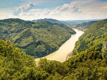 Aerial view of the Danube River winding through the forested Strudengau valley in Austria, with green hills and a small settlement along the shore.