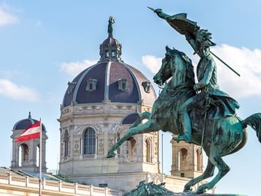 Bronzene Reiterstatue am Heldenplatz in Wien mit österreichischer Flagge und historischer Palastkuppel im Hintergrund unter blauem Himmel.