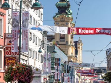 Pedestrian street in Linz with tram tracks, cyclists, and historic church tower with green copper dome. Colorful buildings and hanging flower baskets line the street.