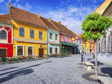 Reihe bunter historischer Gebäude in Szentendre mit Außenbestuhlung auf einem Kopfsteinpflasterplatz unter blauem Himmel mit grünem Baum.