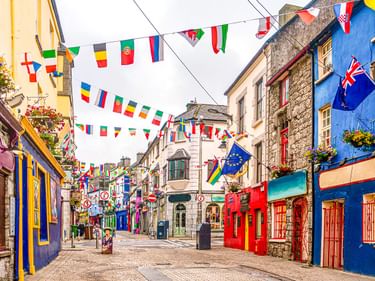 Fußgängerstraße in Galway mit bunten Gebäudefassaden in Blau, Rot, Gelb und Lila. Internationale Flaggen hängen an Schnüren über der Straße.