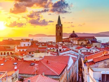 Sonnenuntergang über Algheros Altstadt mit roten Ziegeldächern, hohem Kirchturm und dem Mittelmeer im Hintergrund.