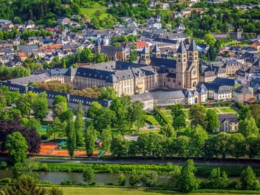 Luftaufnahme der Echternacher Basilika mit zwei Türmen, umgeben von historischen Gebäuden, grünen Parks und Wohngebieten entlang eines Flusses.