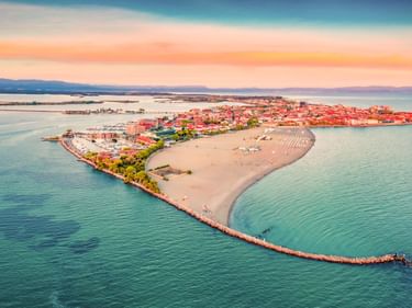 Luftaufnahme der Halbinsel Grado mit rotgedeckten Gebäuden, Sandstrand und türkisfarbener Adria bei Sonnenuntergang.
