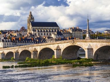 Steinbrücke mit mehreren Bögen über die Loire in Blois, mit Kathedrale und historischen Gebäuden unter dramatischem Wolkenhimmel.