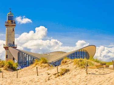 Leuchtturm Warnemünde und das markante wellenförmige Teepott-Gebäude hinter Sanddünen mit Strandgras unter blauem Himmel mit weißen Wolken.