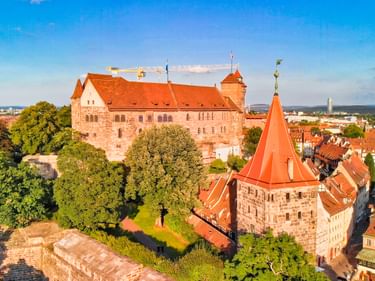 Luftaufnahme der Nürnberger Burg mit orangeroten Dächern und rundem Turm. Mittelalterliche Steingebäude umgeben von grünen Bäumen.