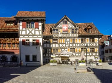 Traditionelle Fachwerkhäuser mit Schweizer Flaggen auf einem sonnigen Stadtplatz in Arbon. Weiße Fassaden mit dunklen Holzbalken und Ziegeldächern.