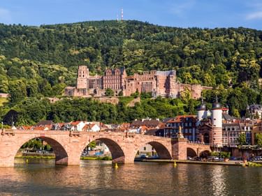 Historische Steinbrücke mit Bögen über den Neckar in Heidelberg, darüber die rote Sandsteinruine des Schlosses auf einem bewaldeten Hang.
