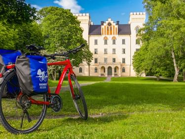 Rotes Fahrrad mit blauen Taschen auf grüner Wiese vor weißem Schloss Börgesund mit Türmen, umgeben von grünen Bäumen unter blauem Himmel.