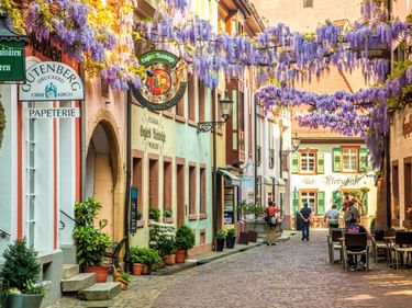 Malerische Kopfsteinpflasterstraße in Freiburgs Altstadt mit bunten historischen Gebäuden und lila blühenden Glyzinien.