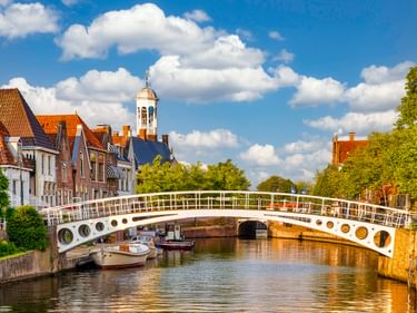 Weiße Fußgängerbrücke mit runden Öffnungen überspannt einen Kanal in Dokkum. Historische Gebäude mit orangefarbenen Ziegeldächern und Kirchturm säumen das Wasser.