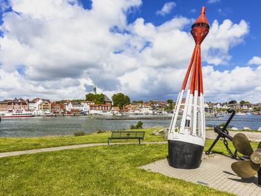 Rot-weiße Navigationsboje als Denkmal mit Schiffspropellern auf grüner Wiese mit Blick auf den Hafen von Kappeln mit historischen Gebäuden und Booten an der Schlei.