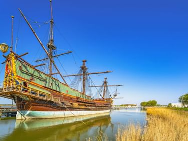 Nachbau des Segelschiffs Batavia mit grün-goldenem Rumpf im Hafen von Lelystad unter blauem Himmel mit ruhigem Wasser und Schilf.