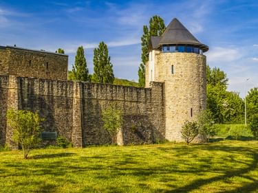 Mittelalterliche Stadtmauer mit rundem Turm in Echternach, Luxemburg. Grüne Wiese im Vordergrund, Bäume umgeben die Befestigung unter blauem Himmel.