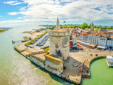 Luftaufnahme des Tour de la Lanterne, ein mittelalterlicher Steinturm im Hafen von La Rochelle, umgeben von Wasser und historischen Gebäuden.