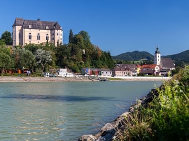 Schloss Greinburg auf einem bewaldeten Hügel über der Donau, mit einem Dorf und einer Kirche darunter, umgeben von grünen Bergen unter blauem Himmel.