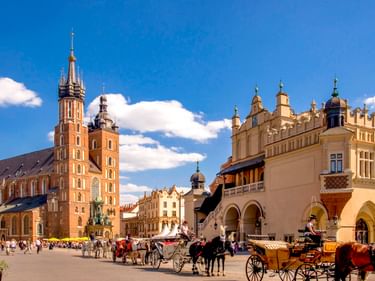 Krakauer Hauptmarkt mit den Zwillingstürmen der Marienkirche und den Renaissance-Tuchhallen. Pferdekutschen und Touristen auf dem Platz.