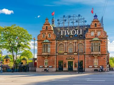 Rotes Backsteingebäude am Eingang des Tivoli mit kunstvoller Architektur und großem beleuchtetem Schild. Dänische Flaggen wehen von Türmen.