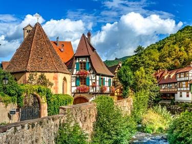 Bunte Fachwerkhäuser entlang der Weiss in Kaysersberg, Frankreich. Steinbrücke und grüne Vegetation säumen den Wasserlauf unter blauem Himmel.