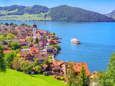 Dorf Beckenried mit Kirchturm am Ufer des Vierwaldstättersees, Raddampfer auf blauem Wasser, grüne Hügel und Berge im Hintergrund.