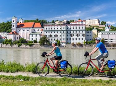 Zwei Radfahrer mit roten Rädern und blauen Taschen fahren entlang der Donau in Passau. Historische Gebäude und Kirche mit orangem Dach am gegenüberliegenden Ufer.